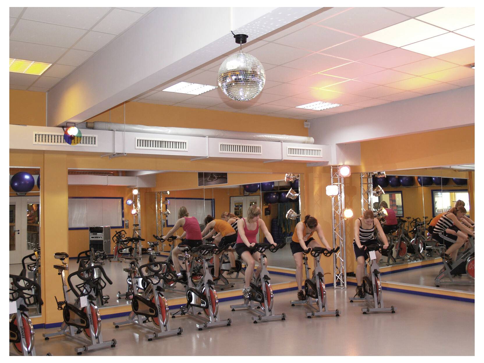 Les gens font de l'exercice sur des vélos stationnaires dans un studio de gym avec miroirs, lumières brillantes et une boule à facettes.