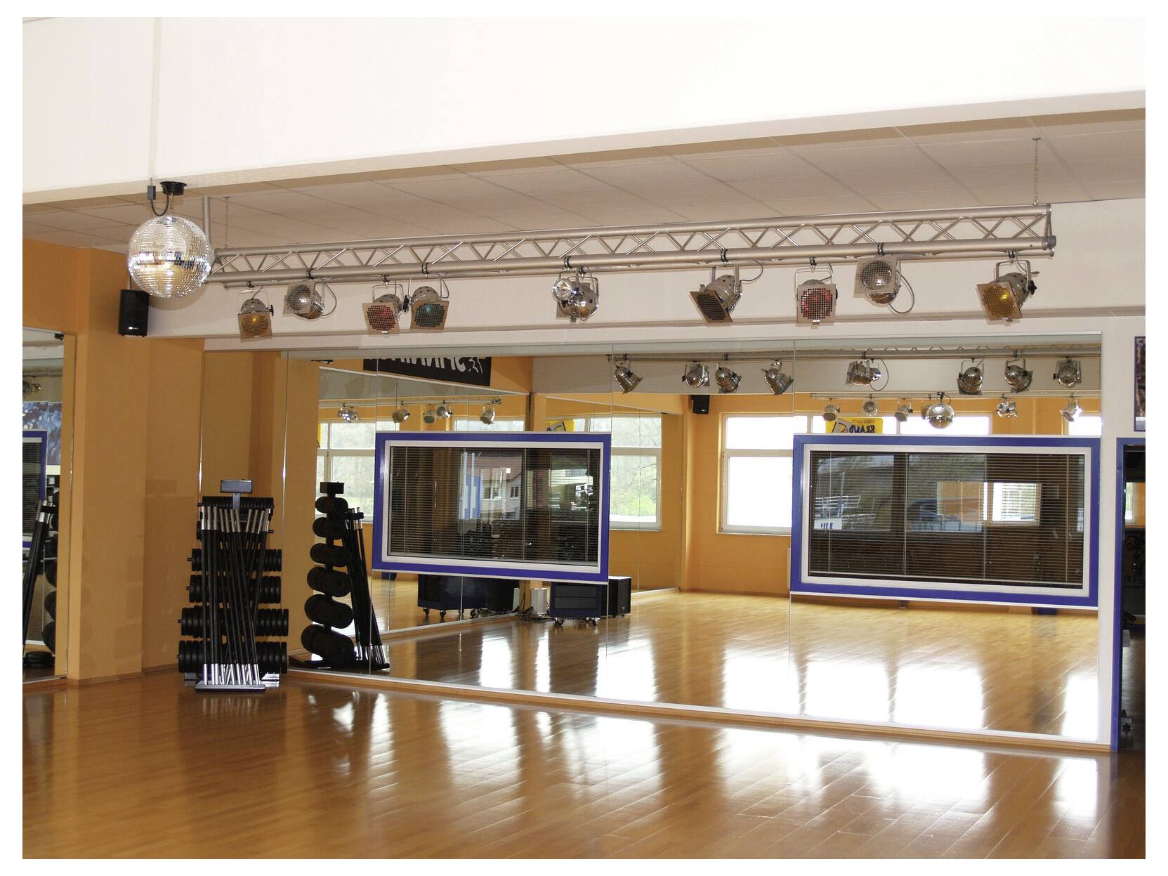 Studio de danse vide avec un mur en miroir, un plancher en bois et des projecteurs de scène montés au plafond ; des marches empilées sont posées à gauche.