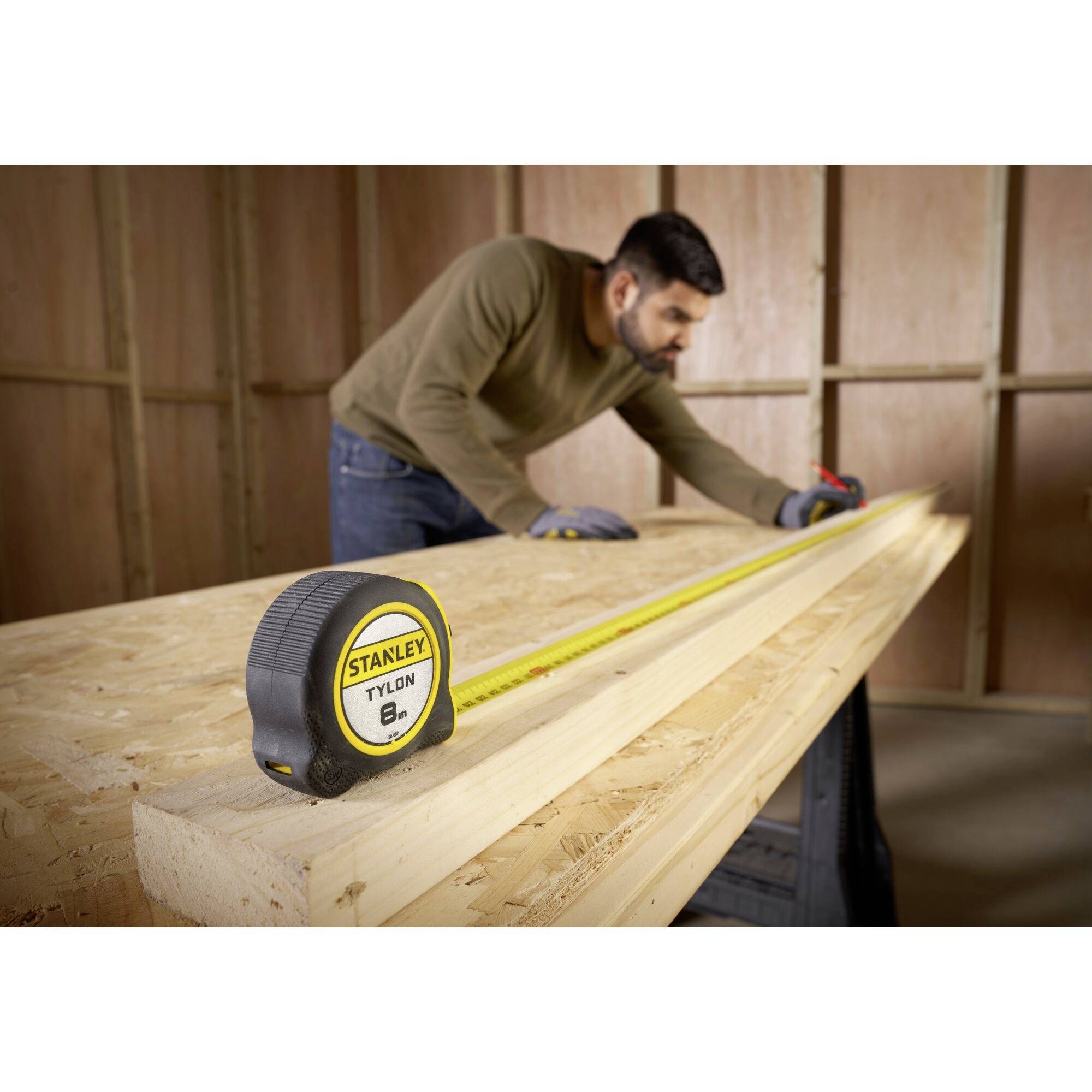 Un homme mesure une longue planche en bois avec un mètre ruban dans un atelier. Au premier plan, le mètre ruban de la marque 'Stanley' est visible.