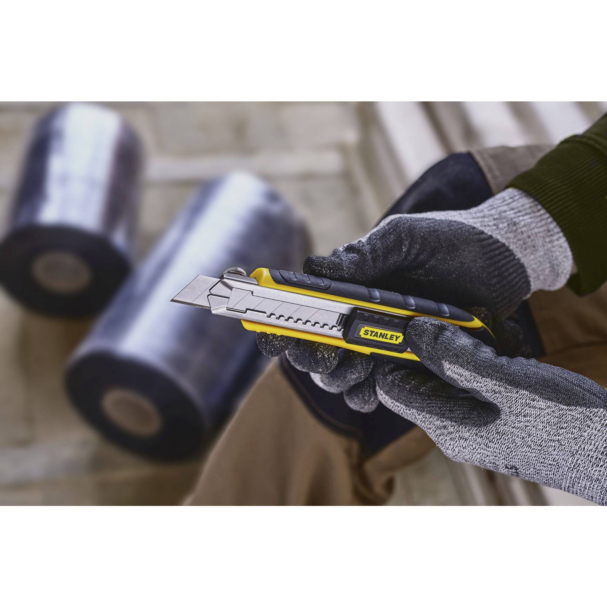 Une personne portant des gants tient un cutter jaune avec la lame sortie. L'arrière-plan est flou et montre de grands rouleaux de film plastique.