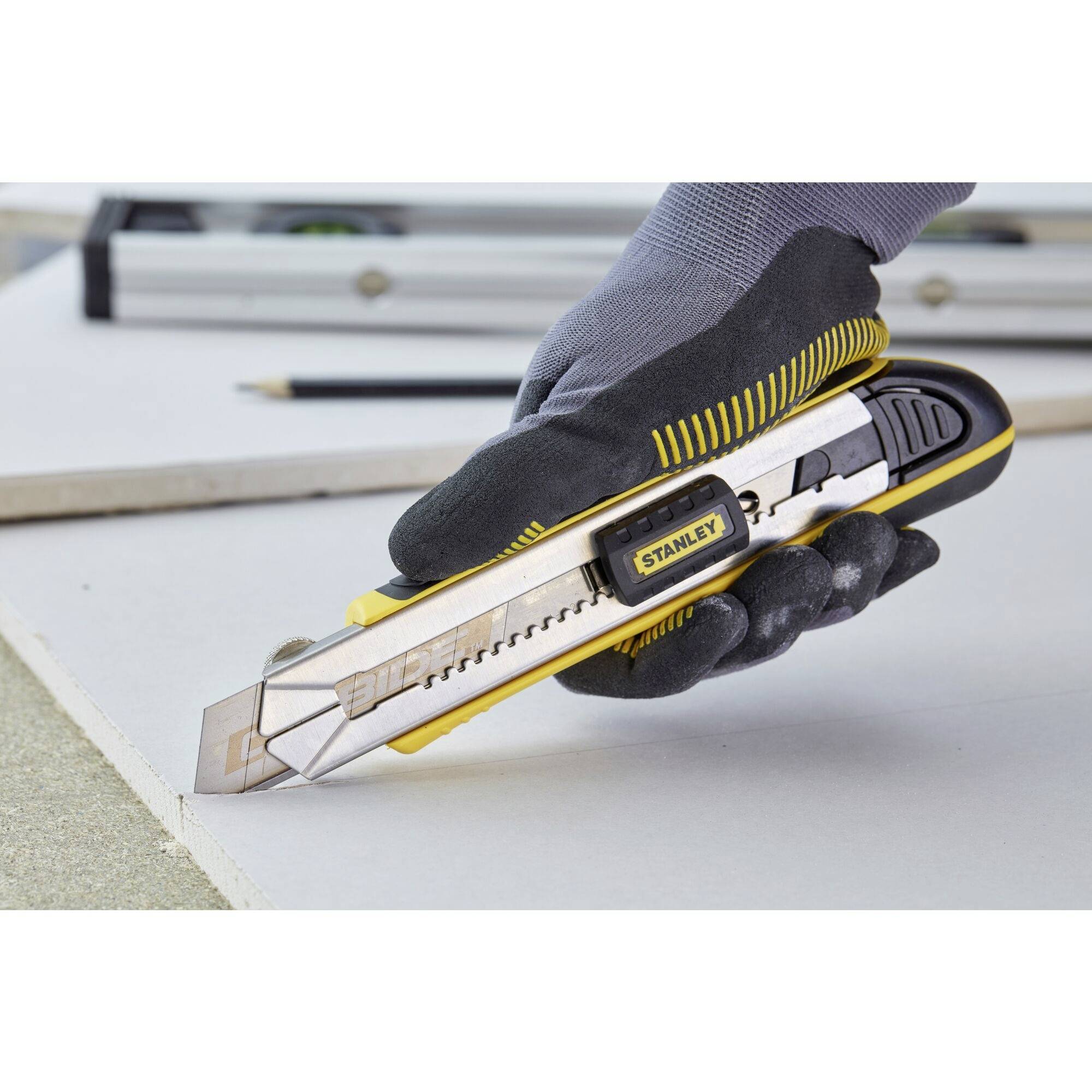 Une main portant un gant de travail tient un cutter jaune sur une surface de travail blanche. Un règle et un crayon sont visibles en arrière-plan.