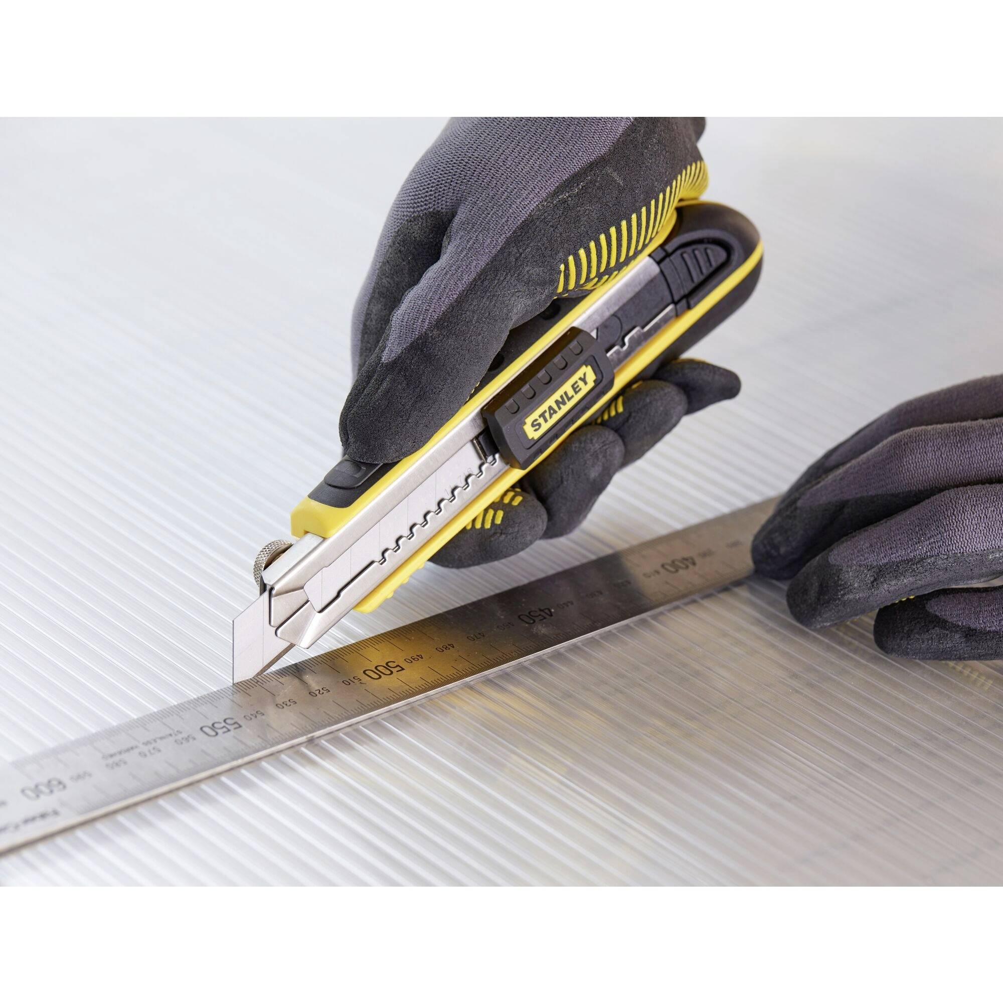 Deux mains gantées découpent le long d'une règle métallique sur une surface texturée à l'aide d'un cutter.