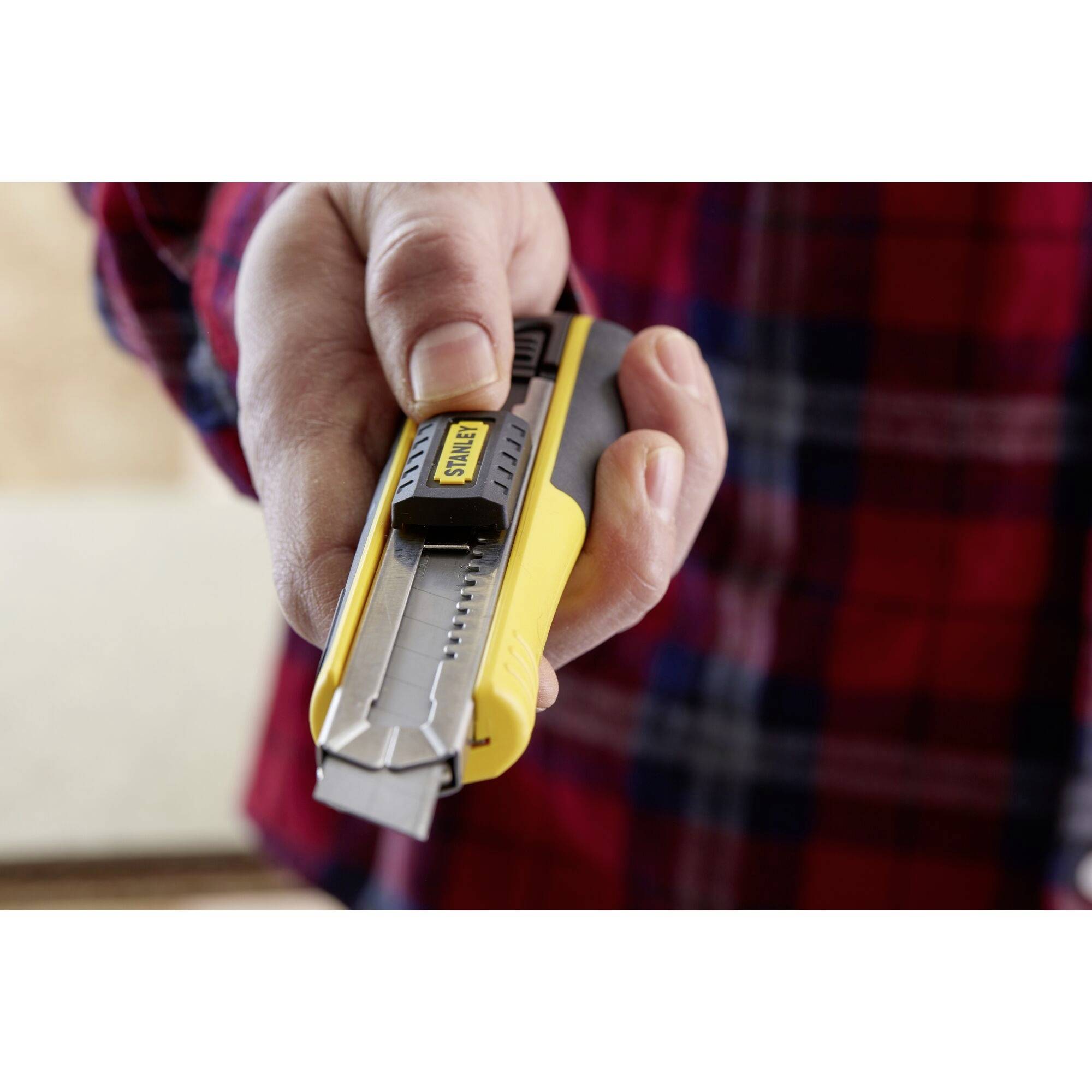 Une main tient un cutter jaune avec une poignée noire, prêt à couper quelque chose. Une personne porte une chemise en flanelle à carreaux.