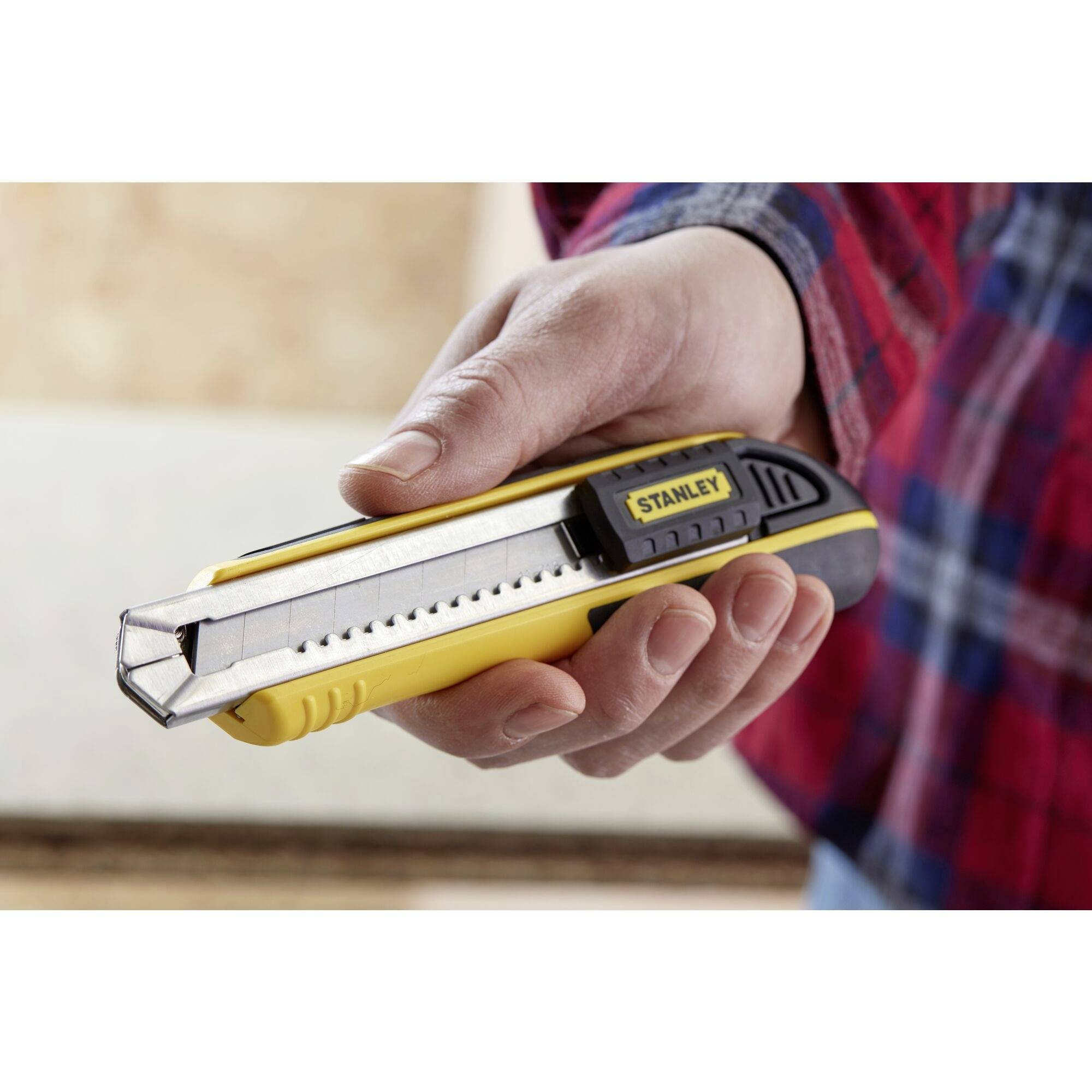 Une main tient un couteau à tapis jaune et noir. La lame est visible. Un plateau en bois est flou en arrière-plan.