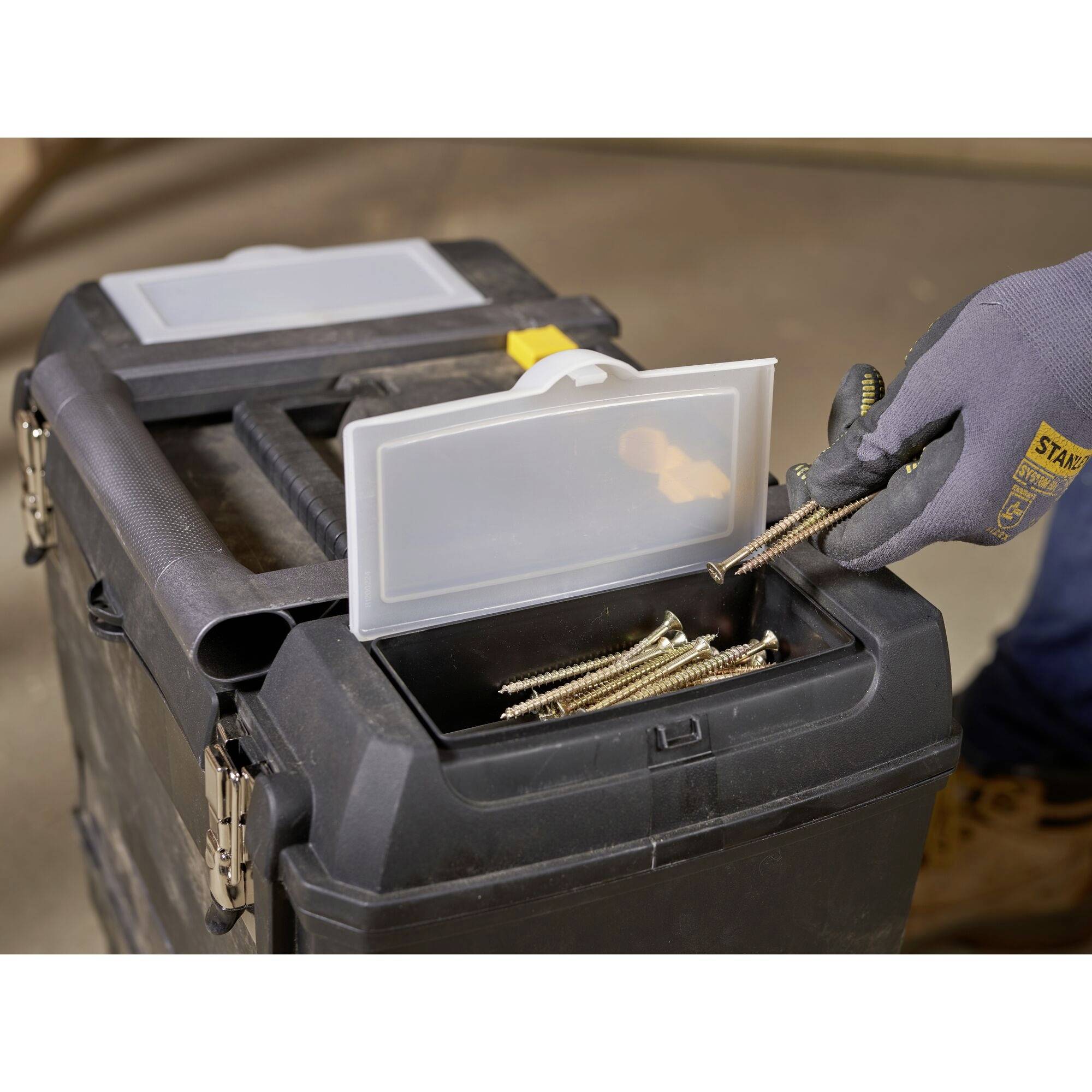 Une personne portant un gant saisit des vis dans un compartiment ouvert d'une boîte à outils. La boîte à outils est partiellement visible.