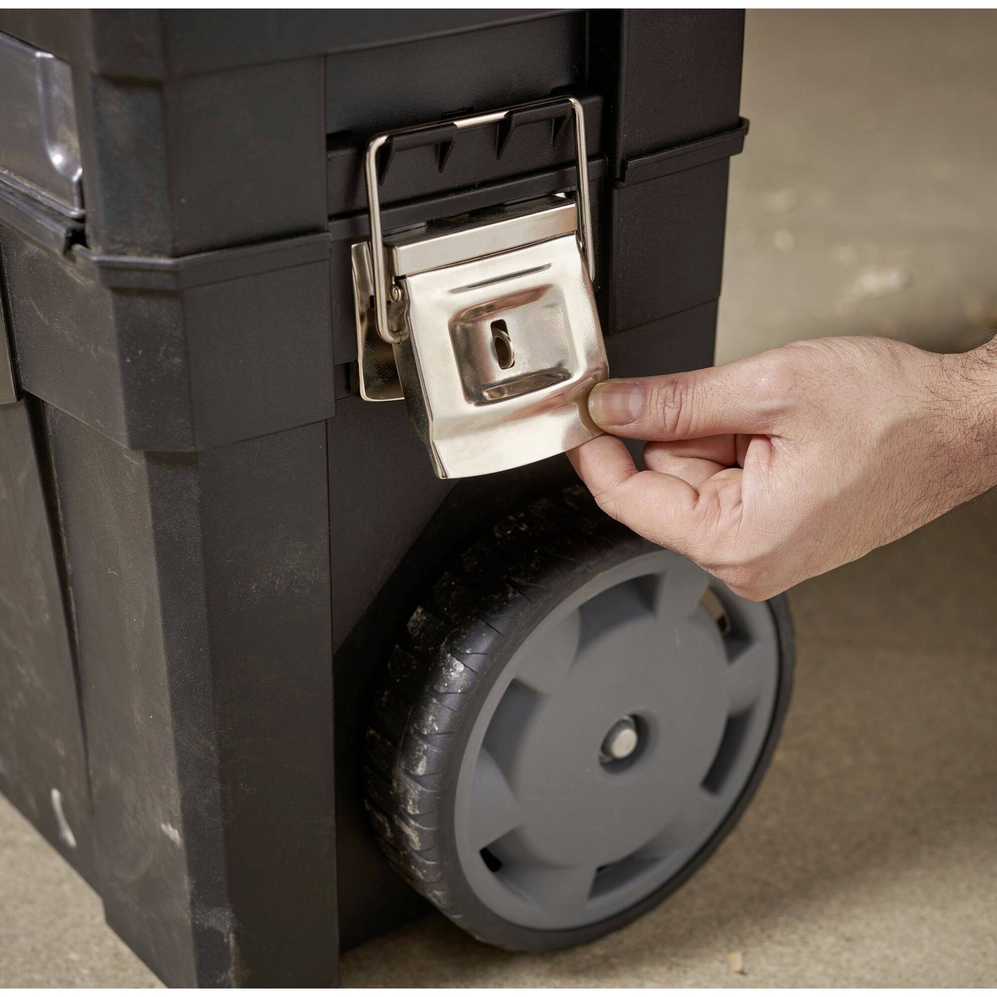 Une main ouvre un verrou métallique sur une valise à roulettes noire avec de grandes roues posée au sol.