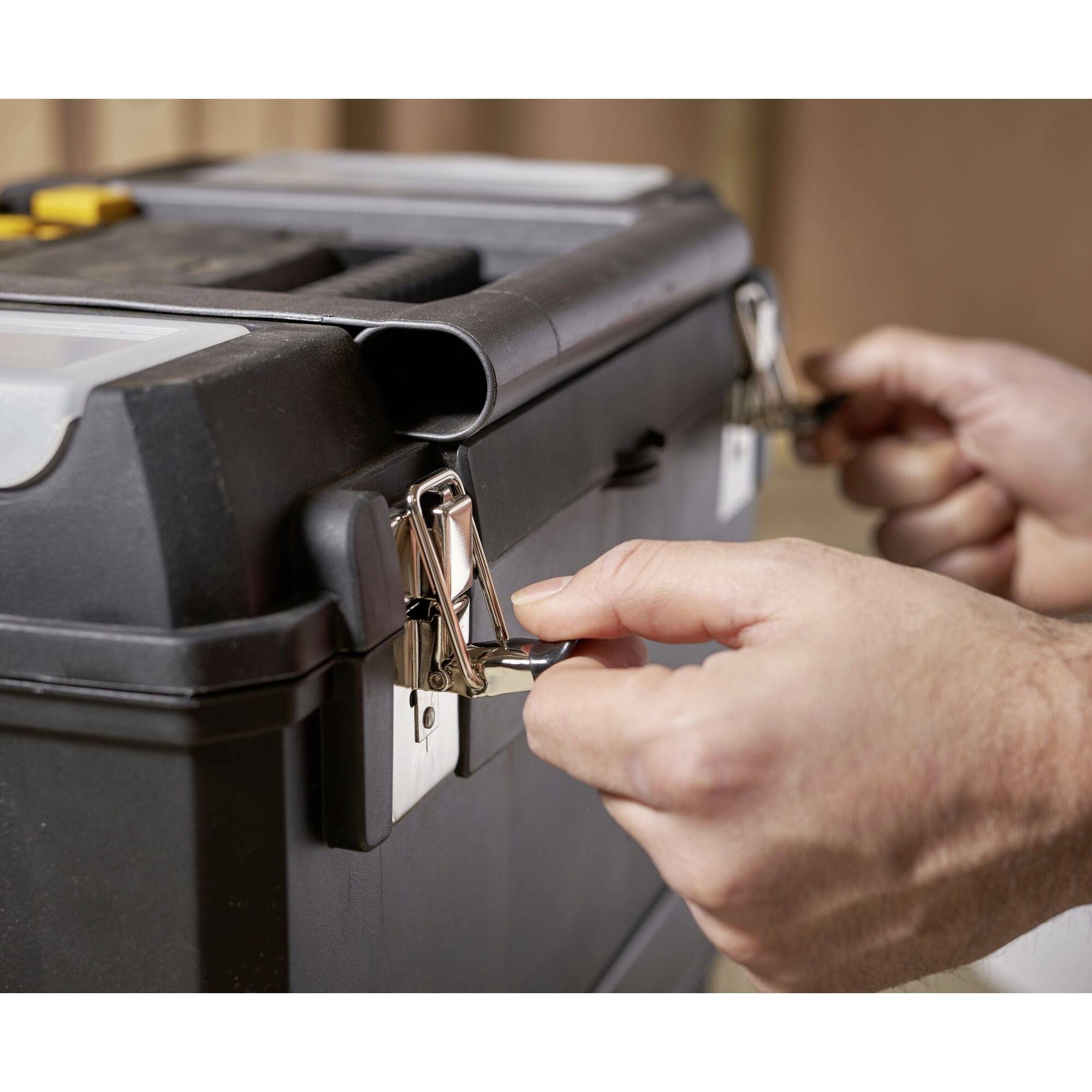 Des mains ouvrent une boîte à outils avec des fermoirs métalliques. La boîte est noire et se trouve dans une pièce au fond flou.