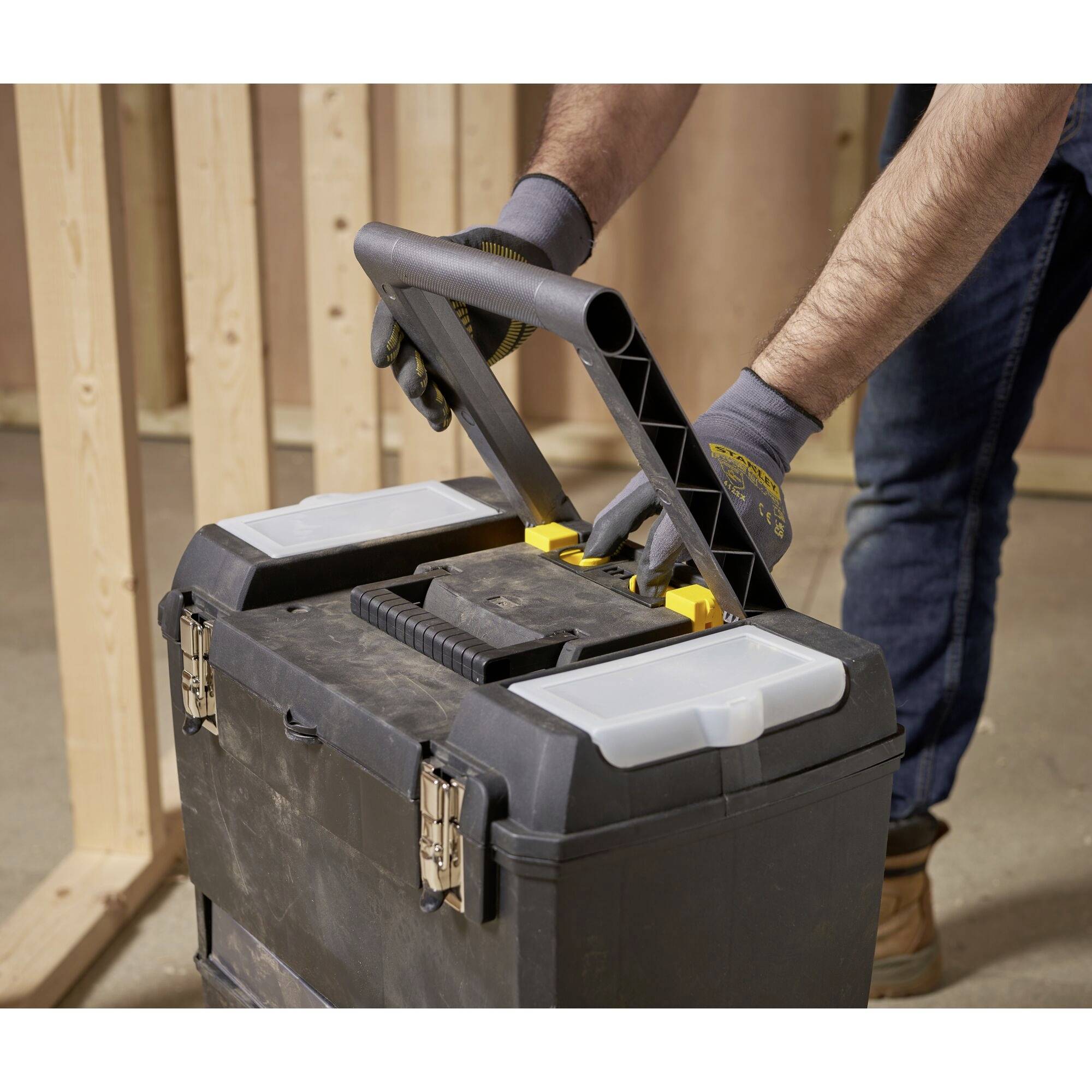 Une personne porte une boîte à outils noire avec des éléments jaunes, debout dans une pièce à ossature en bois non finie. Elle porte des gants de travail.