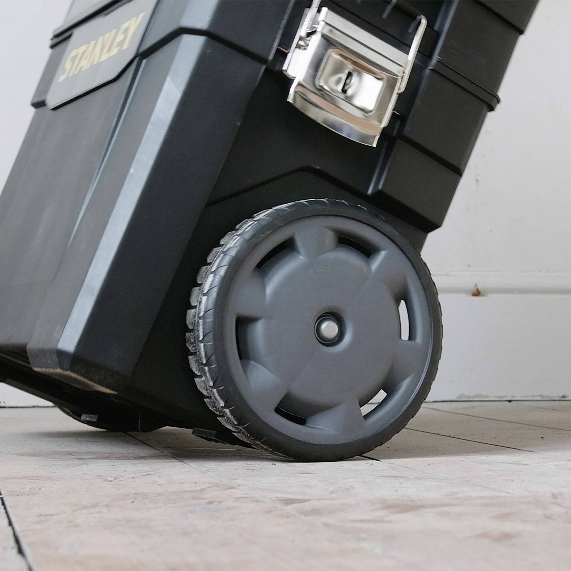 Boîte à outils noire avec roulettes grises et fermeture argentée sur un sol carrelé.