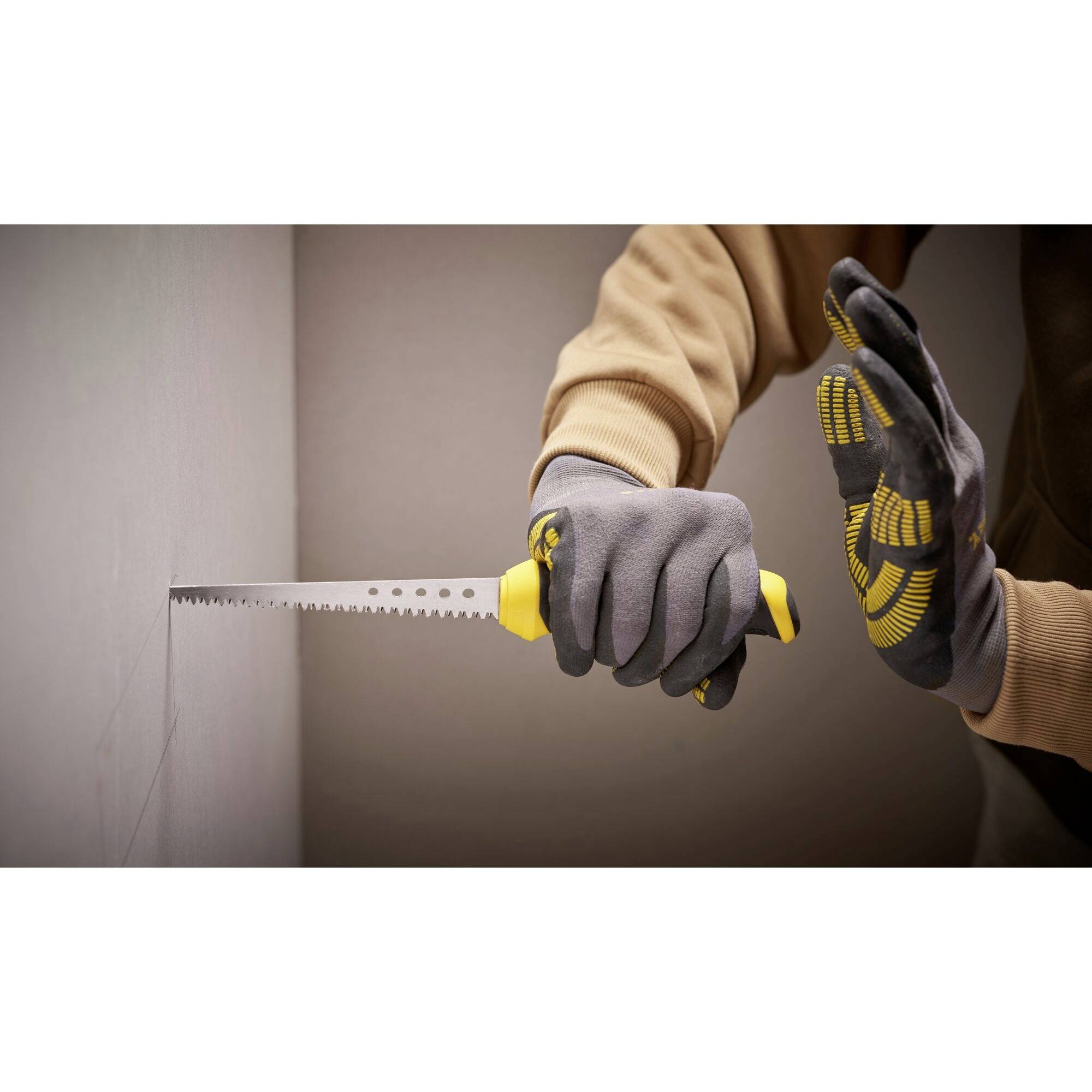 Une personne coupe un trou dans un mur à l'aide d'une scie à main. Elle porte des gants gris.