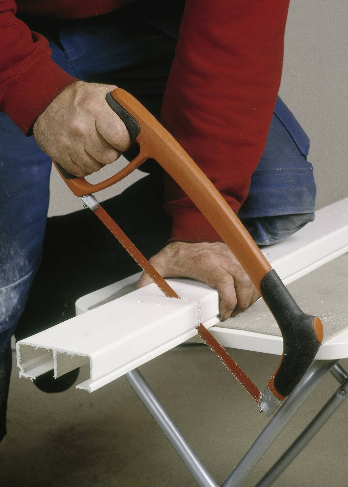 Une personne coupe un tube en plastique blanc avec une scie à métaux sur une table. La personne porte des manches rouges et un jean bleu.
