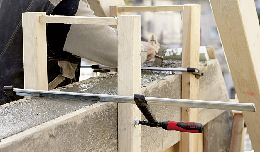 Une personne travaille un coffrage en bois sur un chantier. Un serre-joint maintient le coffrage en place pendant le coulage du béton.