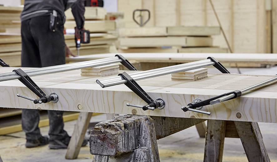 Une personne travaille dans un atelier, pose des serre-joints sur des planches de bois collées afin de les maintenir en place. À l'arrière-plan, différentes pièces de bois sont visibles.