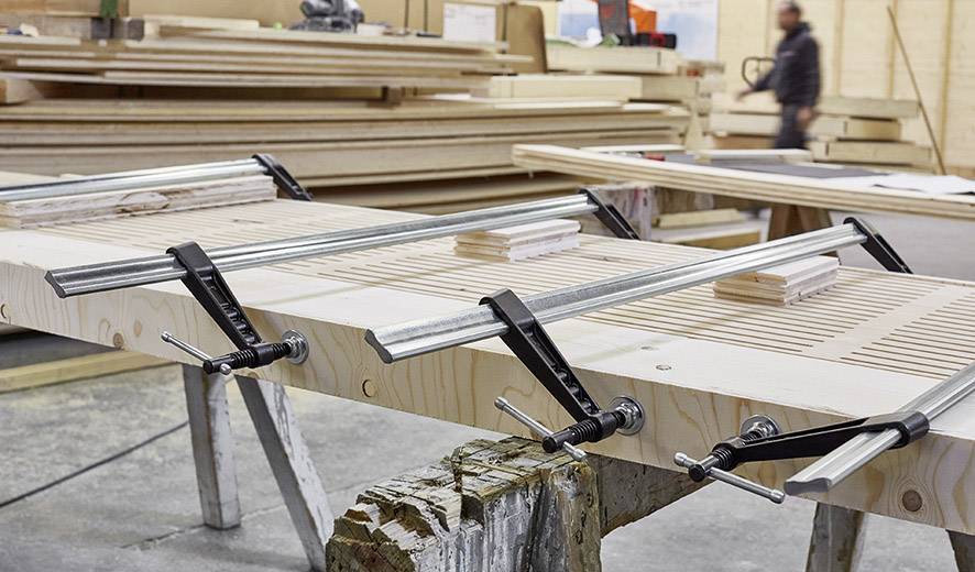 Un plateau en bois est fixé avec des serre-joints métalliques dans un atelier. En arrière-plan, on aperçoit des planches de bois empilées et une personne floue.