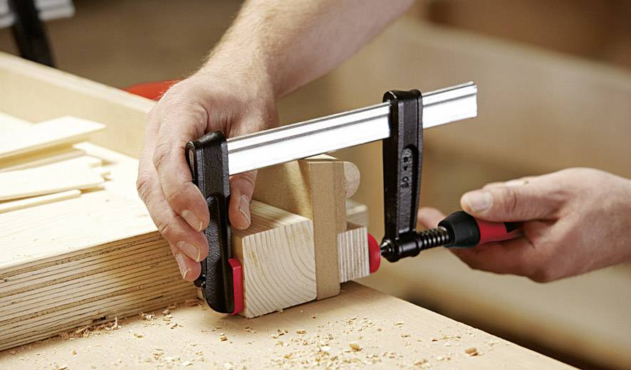 Une personne utilise un serre-joint noir pour fixer des morceaux de bois sur un établi. Des copeaux de bois sont visibles.
