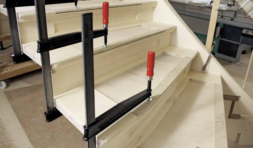 Un escalier en bois mi-construit maintenu par deux serre-joints qui fixent les marches. Des outils et des machines sont visibles en arrière-plan.