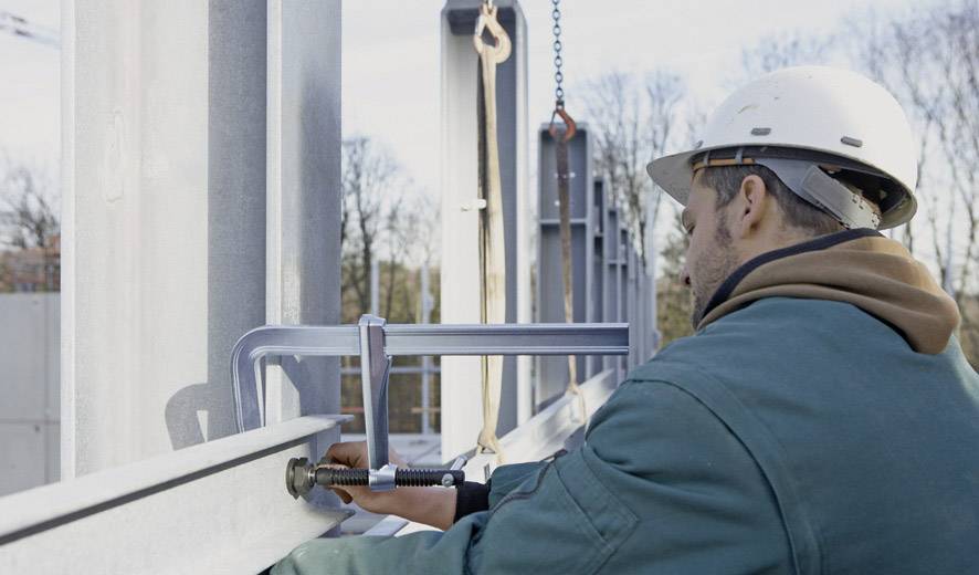 Un ouvrier portant un casque de chantier fixe un élément de poutre métallique sur un chantier à l'aide d'un serre-joint. En arrière-plan, on aperçoit un paysage boisé.