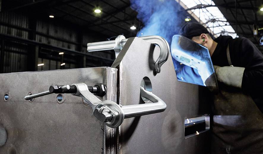Un soudeur travaille sur une grande pièce métallique dans un hall d'usine. Des étincelles bleues jaillissent, et des serre-joints sont installés pour maintenir la stabilité.