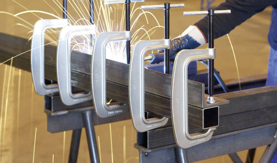 Un ouvrier soude des profilés métalliques, des étincelles jaillissant autour de lui. Plusieurs serre-joints C maintiennent les profilés stables sur un châssis.