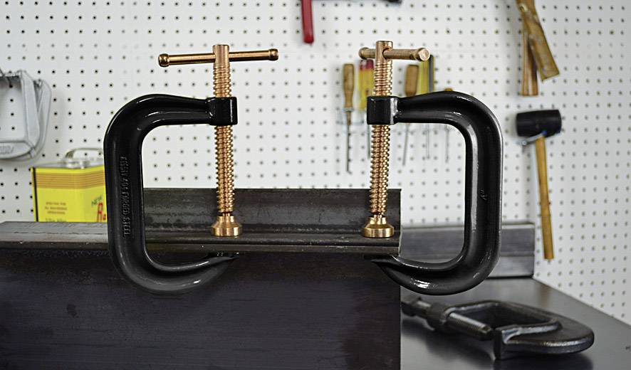 Des étaux de serrage maintiennent une pièce métallique dans un atelier. L'arrière-plan montre des outils accrochés au mur.