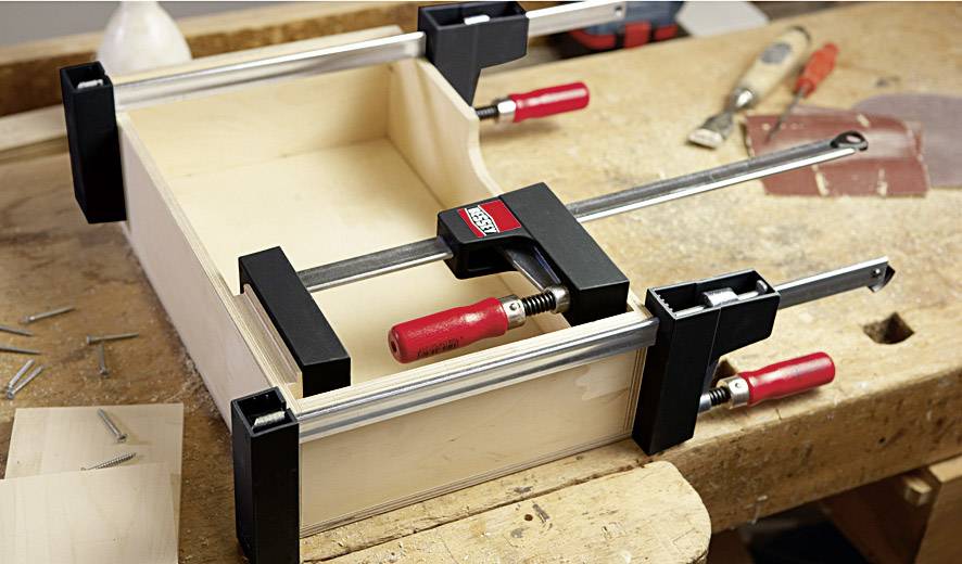 Une boîte en bois est sécurisée avec quatre pinces et travaillée sur un établi. Sur l'établi se trouvent des outils et des clous.