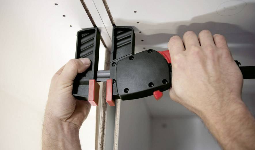 Une personne fixe deux planches à l'aide d'une grande pince rouge. Les mains maintiennent fermement la pince sur les planches en bois.