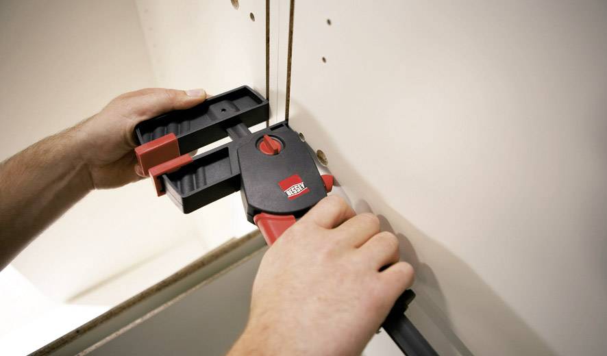 Une personne utilise un serre-joint pour découper un trou rectangulaire dans une cloison en plaque de plâtre.