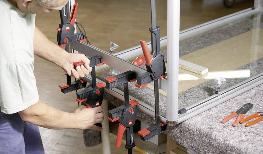 Une personne fixe une pièce métallique à un cadre de table à l'aide de plusieurs serre-joints pour la stabiliser. Des outils sont prêts à être utilisés.