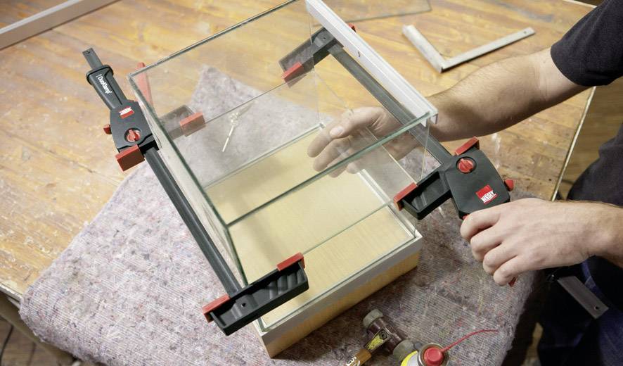 Une personne installe un aquarium en verre à l'aide de pinces de serrage sur une table en bois, présentant un projet DIY dans un environnement d'atelier.