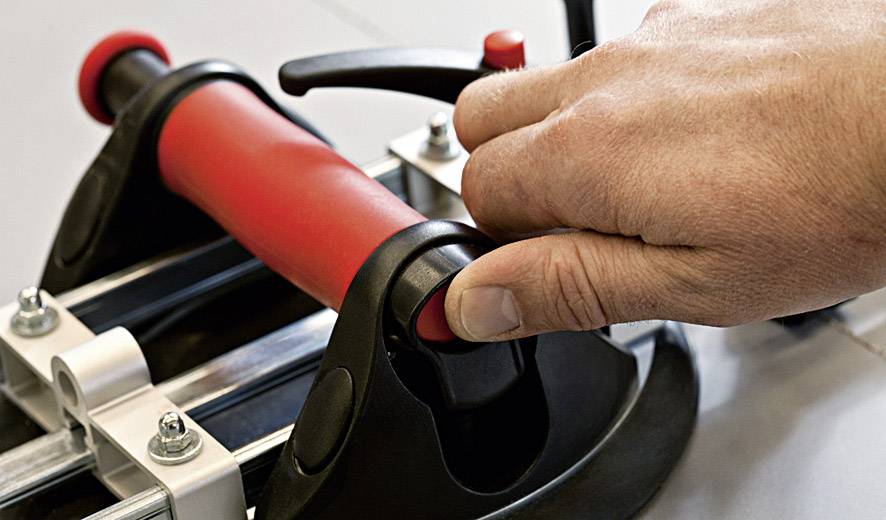 Une main actionne le levier d'un coupe-carrelage à poignée rouge, prête à couper une dalle.