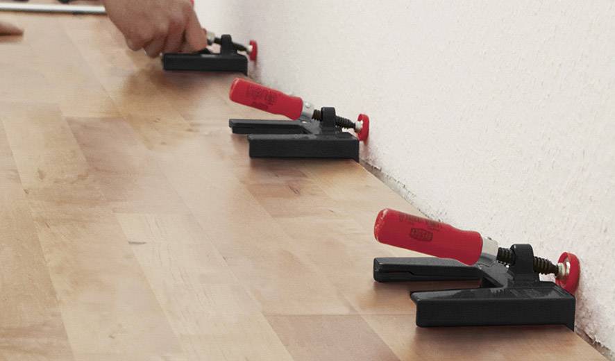 Une main fixe des lames de parquet avec des cales en plastique rouge contre un mur, afin de garantir des espacements uniformes lors de la pose d'un plancher en bois.