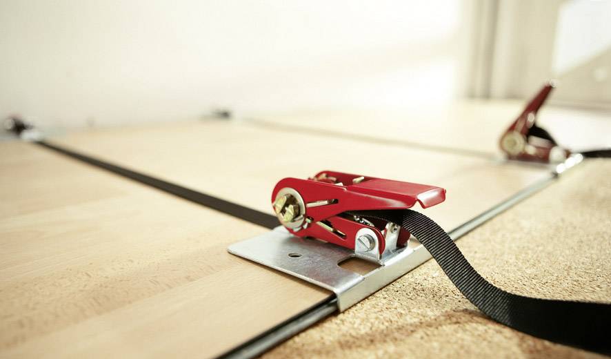 Des sangles à cliquet rouges fixent une planche de bois sur une table.