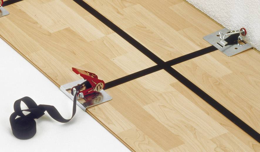 Des sangles et des pinces maintiennent un panneau en bois sur un sol.