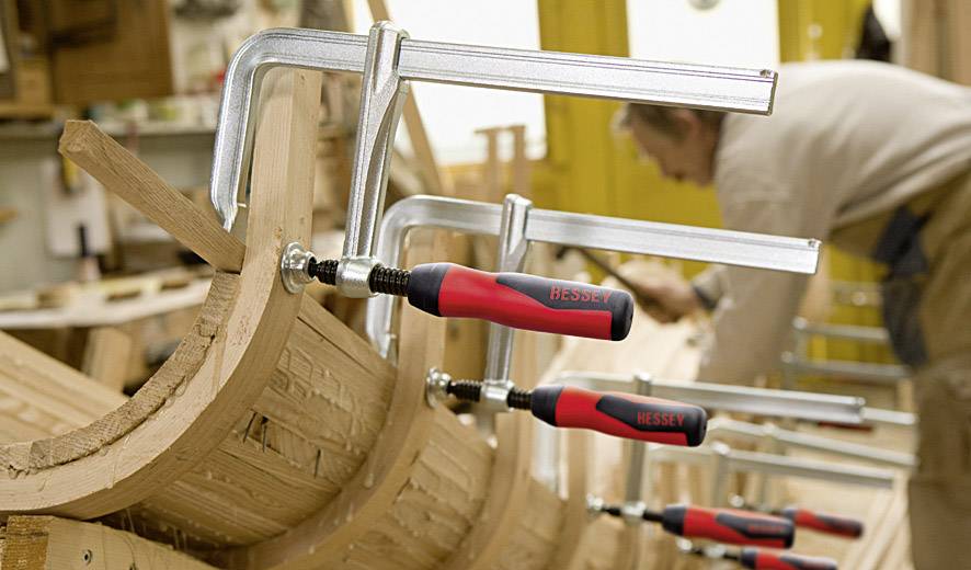 Travail du bois dans l'atelier : Plusieurs serre-joints maintiennent des pièces de bois courbées. Un artisan travaille concentré en arrière-plan.