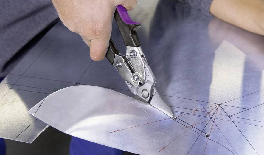Une personne coupe une tôle avec une cisaille métallique, des repères visibles sur le métal indiquent les lignes de coupe.
