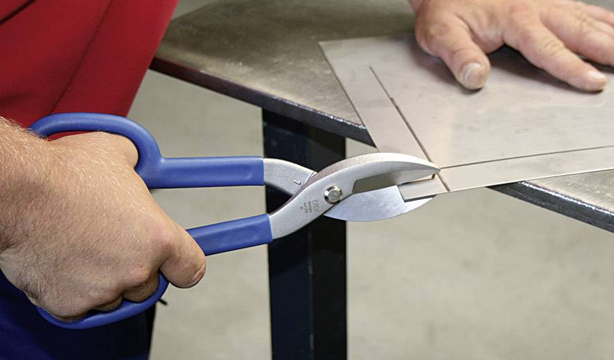 Un homme coupe un morceau de tôle avec des ciseaux bleus en métal au bord d'une table.