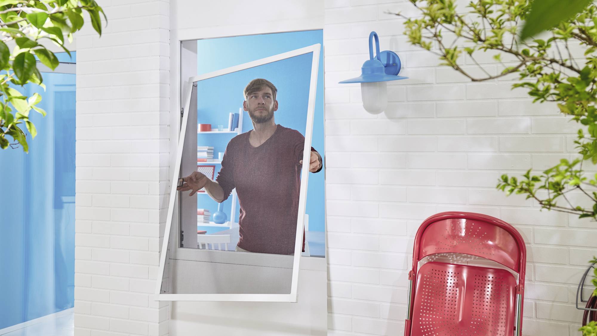 Une personne installe un cadre de fenêtre blanc dans un bâtiment en briques blanches. Au premier plan, une chaise rouge.