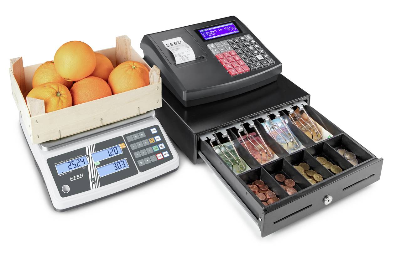 Une caisse enregistreuse électronique avec un compartiment à monnaie ouvert montre différentes pièces et billets. À côté se trouve une balance numérique avec des oranges dans une boîte en bois.