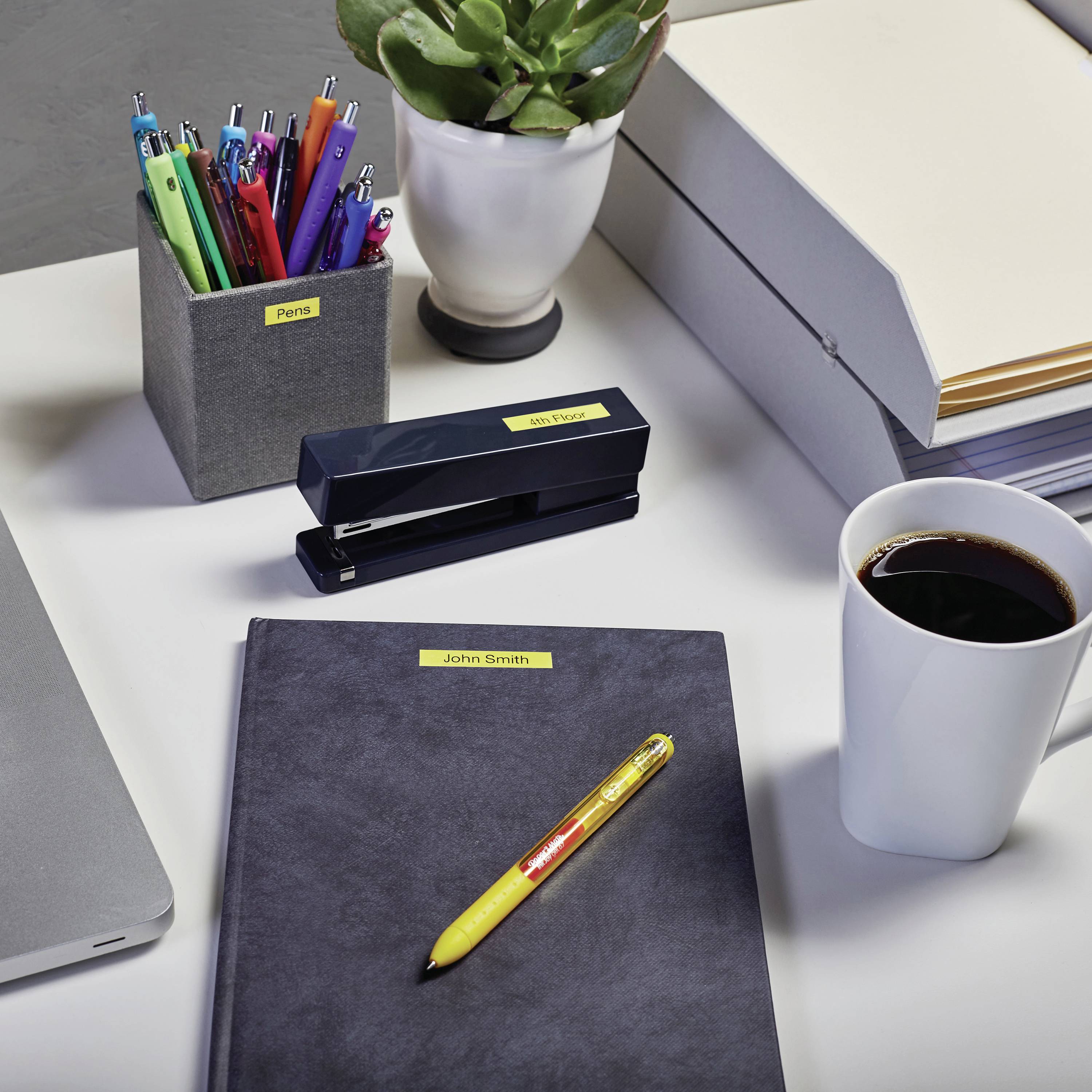Bureau avec un ordinateur portable, des stylos, un distributeur d'agrafes, une plante, une pile de classeurs, un carnet de notes et une tasse de café.