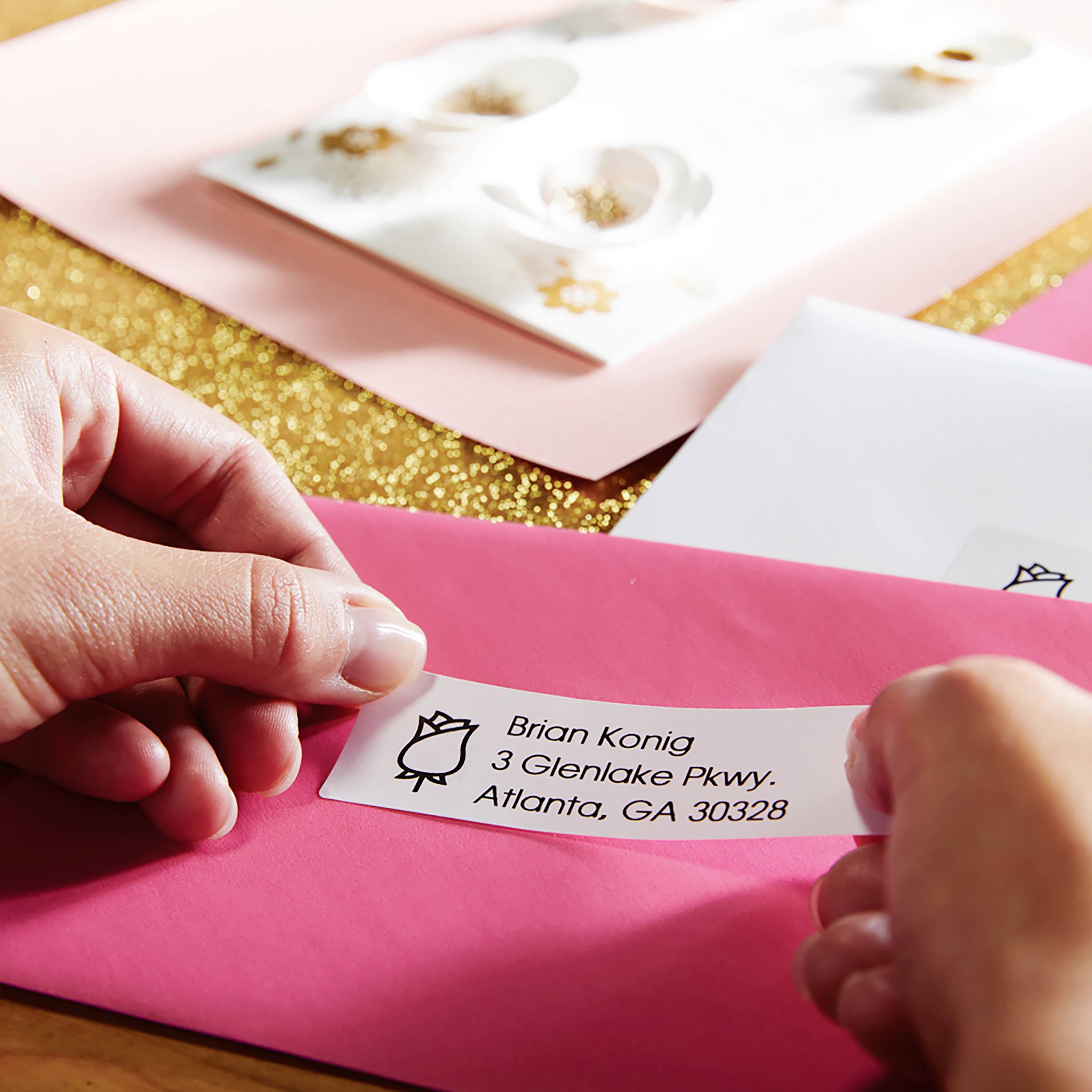 Une main colle une étiquette d'adresse sur une enveloppe rose. L'arrière-plan montre d'autres enveloppes et une surface dorée.
