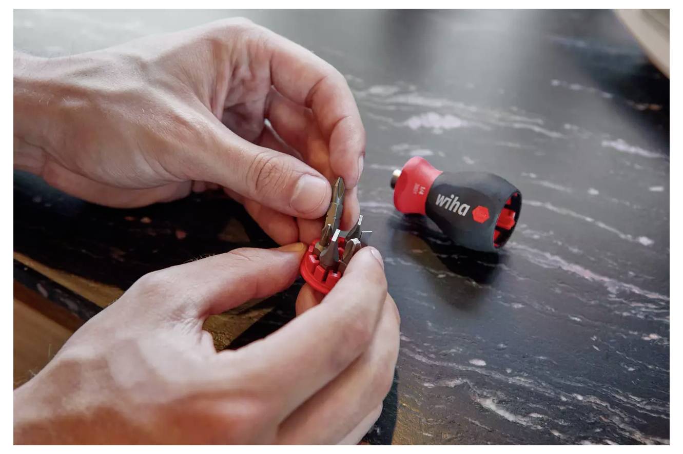 Les mains ajustent un porte-outil rouge avec plusieurs embouts interchangeables ; un petit manche de tournevis noir et rouge repose à proximité sur une surface sombre.