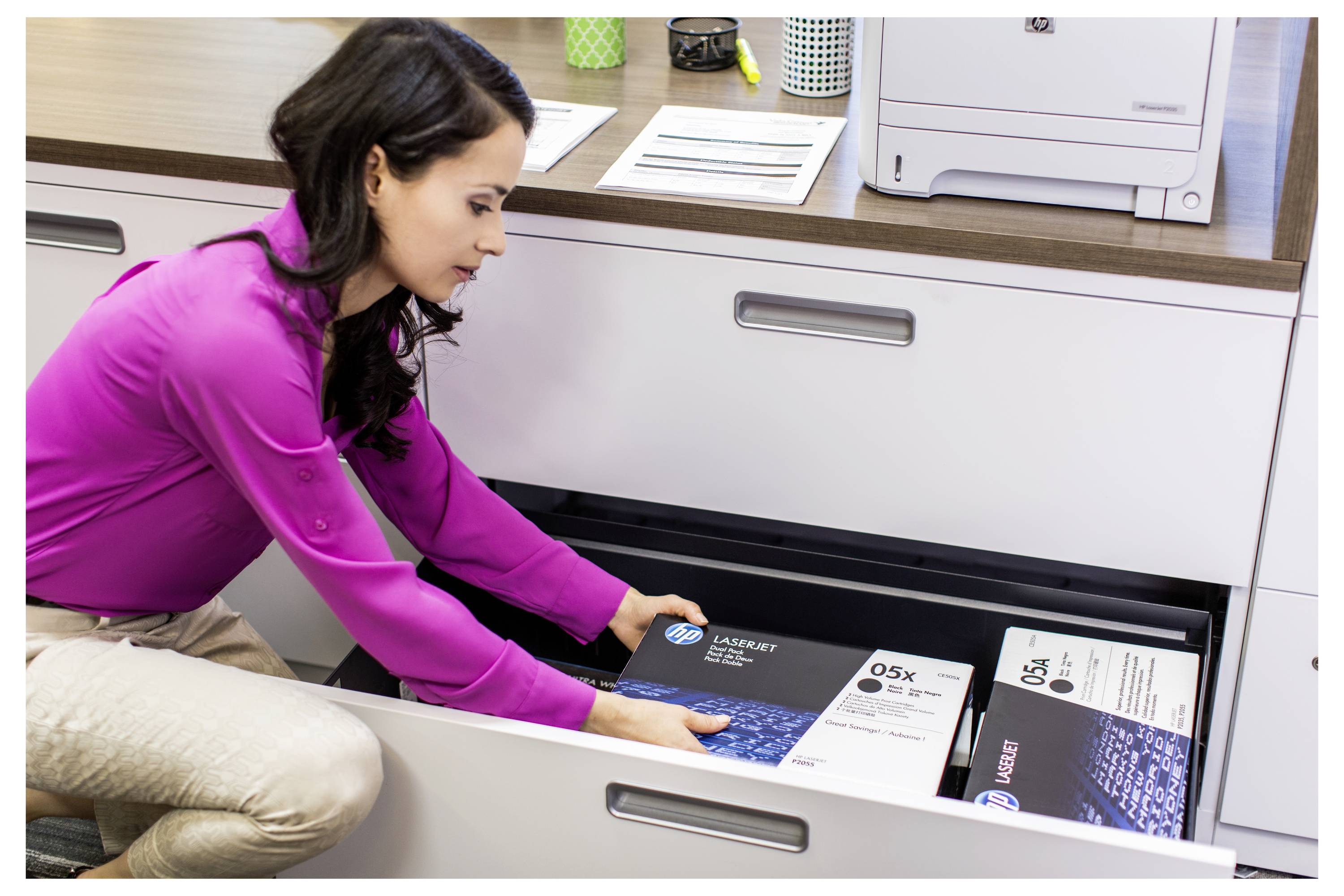 Femme vêtue d'un chemisier violet s'agenouille et ouvre un tiroir de classement, organisant des boîtes de documents dans un bureau ou une salle de photocopie.