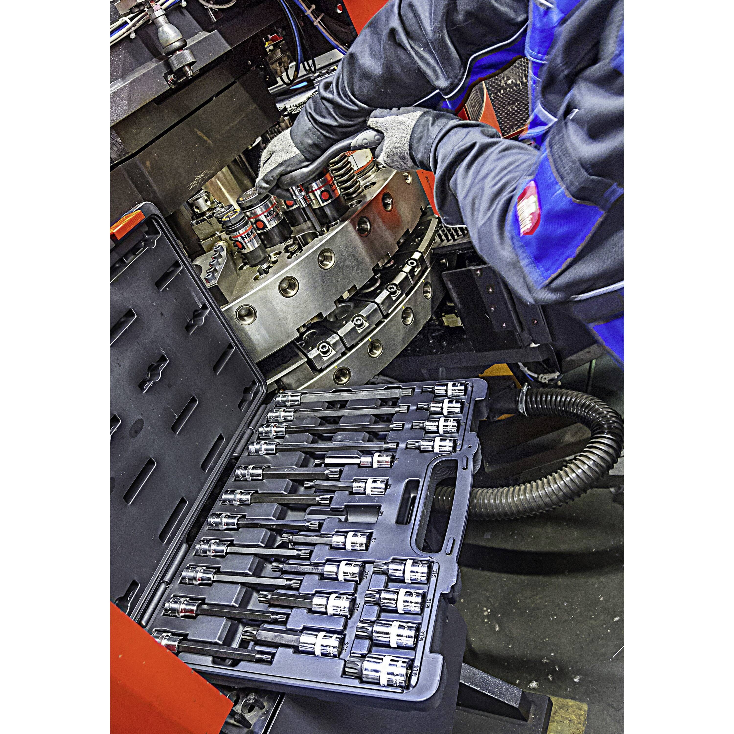 Une personne portant des gants et des vêtements de protection assemble des composants d'outils sur une machine, avec une boîte à outils ouverte montrant plusieurs clés à douille.