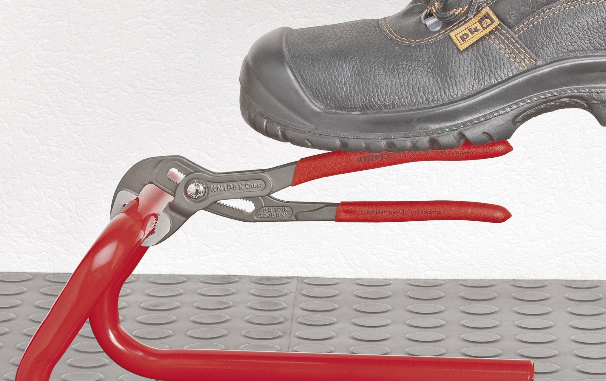 Une chaussure est posée sur une pince rouge qui tient un tuyau rouge. La pince est placée sur une surface grise structurée.