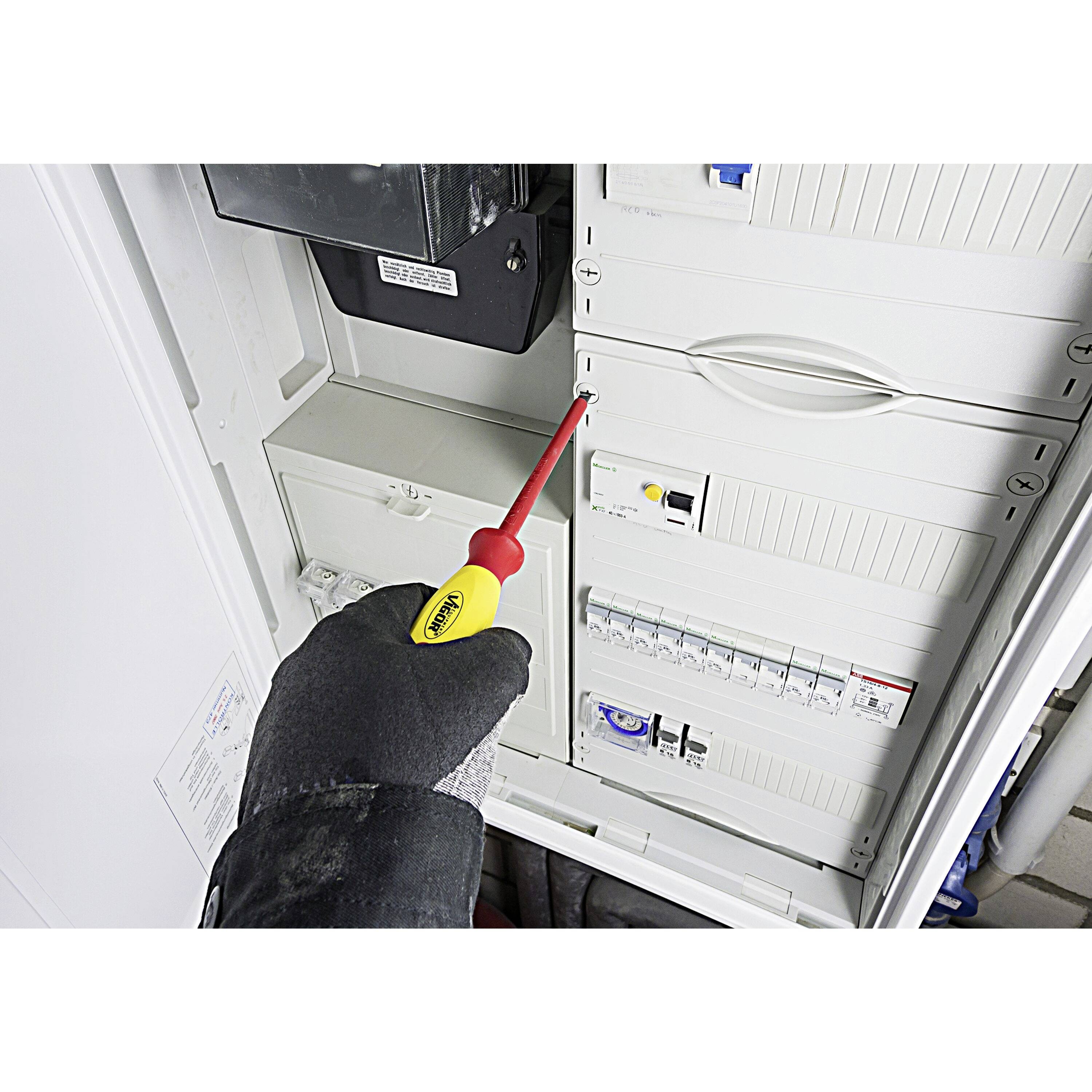 Une personne portant un gant utilise un tournevis jaune et rouge sur un tableau électrique avec plusieurs interrupteurs et composants visibles.