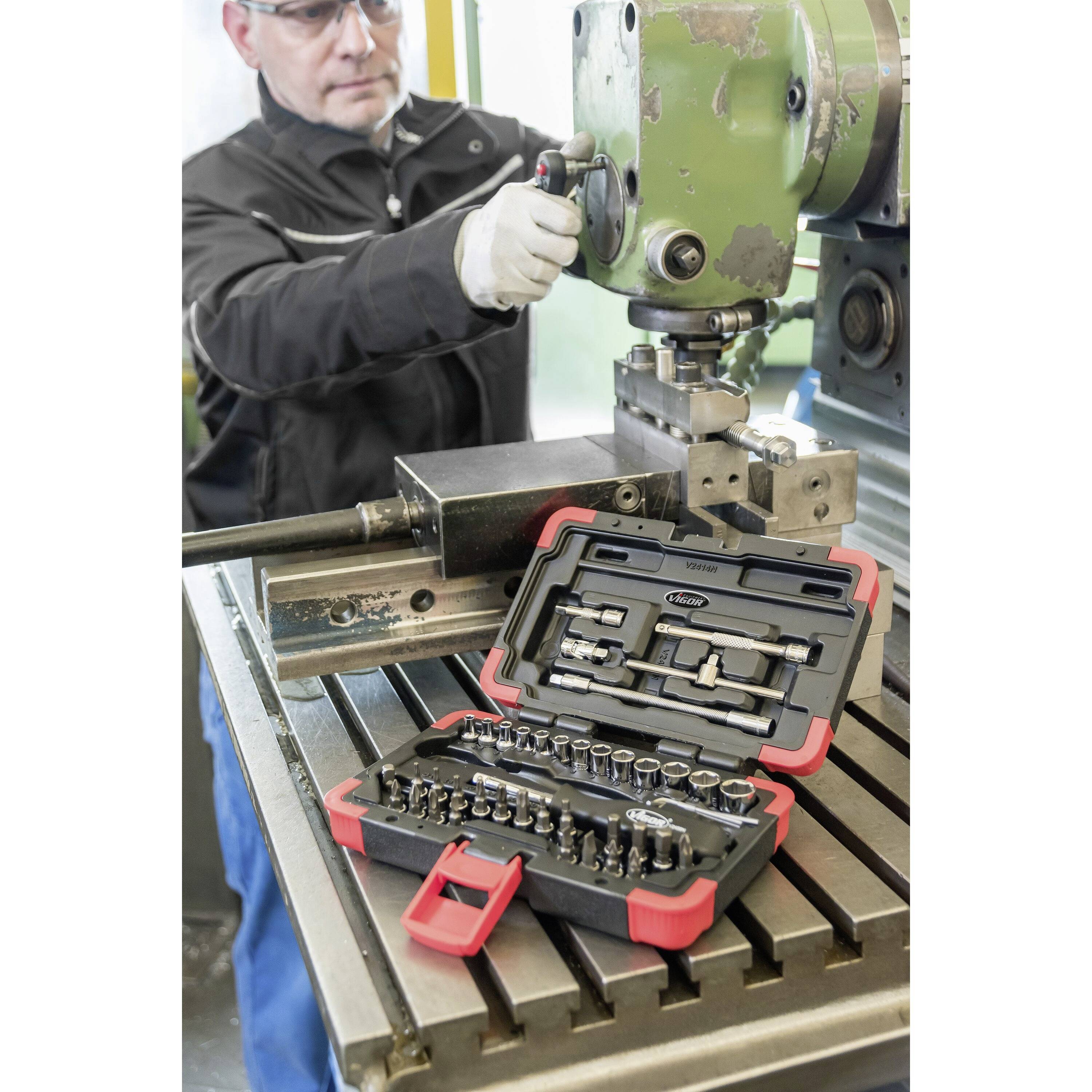 Un artisan qui travaille sur une machine, tandis qu'un coffret d'outils ouvert avec des clés à douille se trouve au premier plan sur une table.