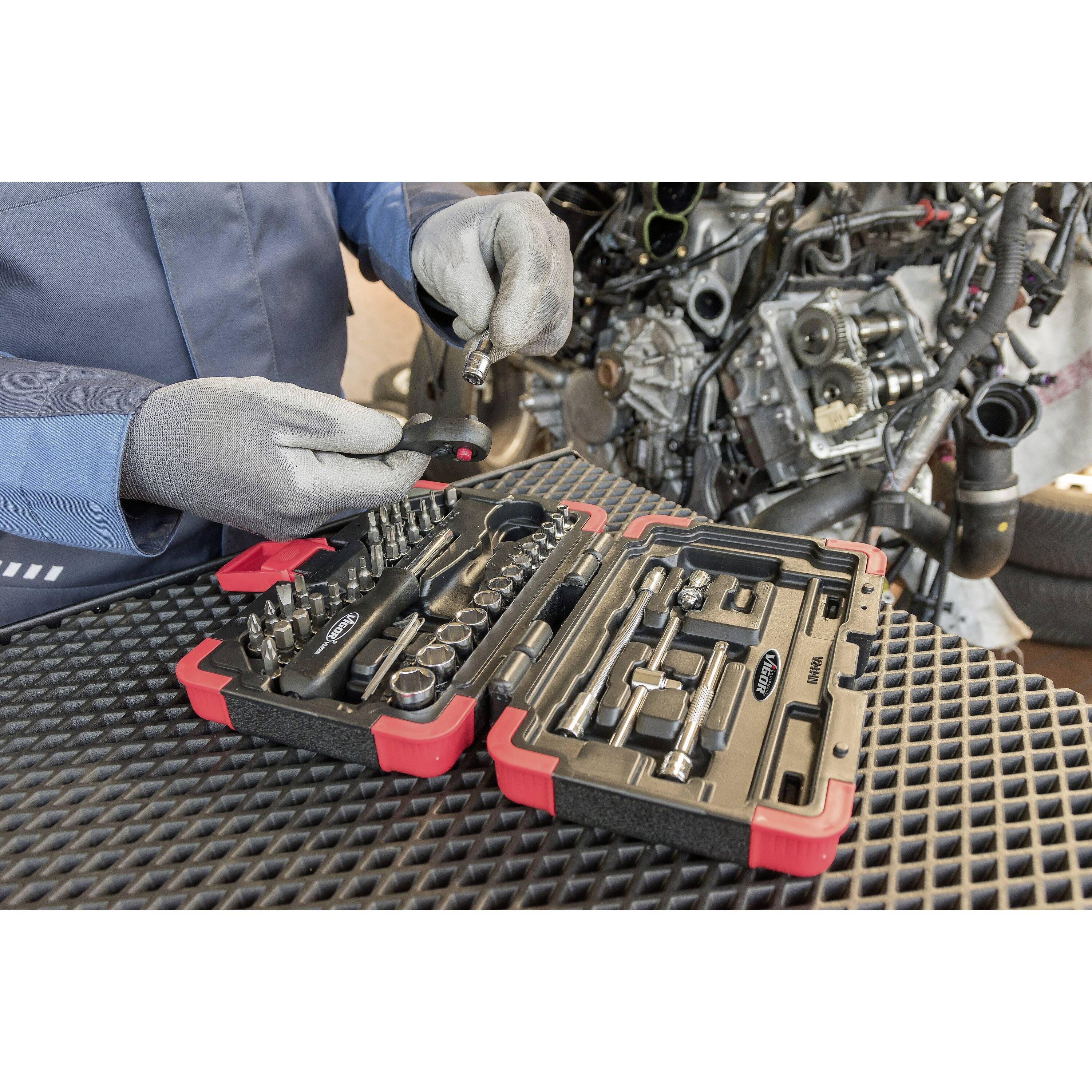 Une personne en vêtements de travail ouvre une boîte à outils contenant diverses clés à douille et travaille sur un moteur posé sur un établi.