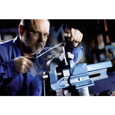 Un homme travaille une pièce métallique dans un étau avec une lime. Il porte des vêtements de travail bleus et des lunettes de protection dans un atelier.