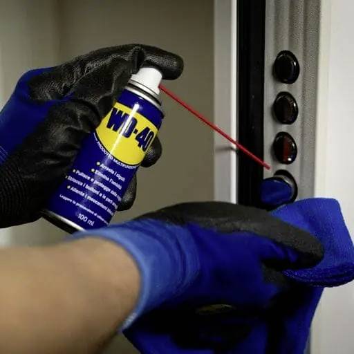 Une paire de mains gantées de bleu vaporise du WD-40 sur les charnières d'une porte pour la lubrifier et réduire les grincements.