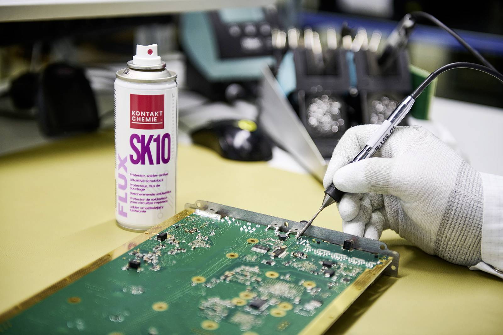 Une main gantée soude un circuit imprimé vert. Une boîte portant l'inscription 'KONTAKT CHEMIE SK10' est posée à côté.